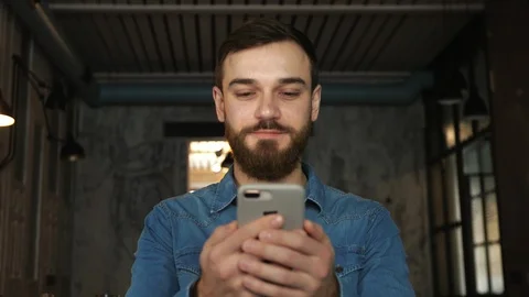 Portrait of a young man with a beard in a cafe. Blue shirt Man writes a message Vídeos de archivo 100604456