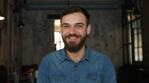 Portrait of a young man with a beard in a cafe. Blue shirt. Smile Stockbeeldmateriaal 100604910