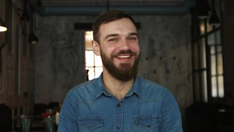 Portrait of a young man with a beard in a cafe. Blue shirt. Smile Vídeos de archivo 100605381