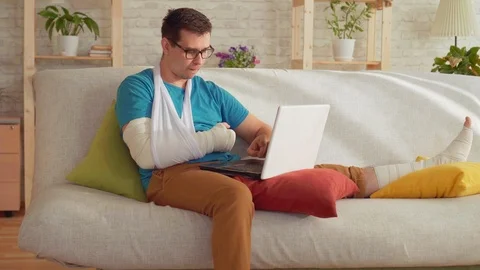 Portrait of a young man with a broken arm and leg sitting on the couch and uses Stock-Footage 106778496