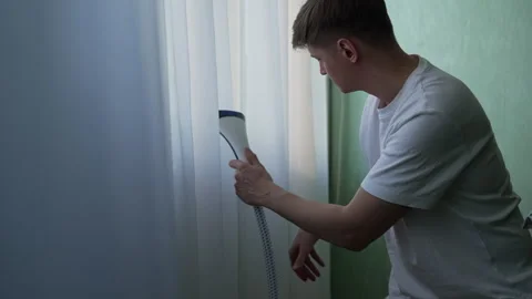 Portrait of young man cleaning steaming wrinkled white curtains near wall Stock Footage 304883464