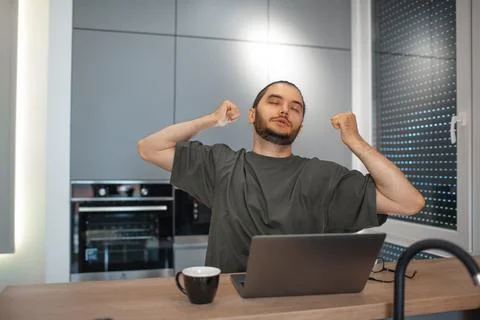 Portrait of young man stretched while working home at laptop on kitchen. Bu.. Stock Photos