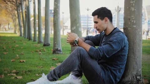 Portrait of young man using smartphone and sitting near tree in warm autumn day, Stock Footage 118194240