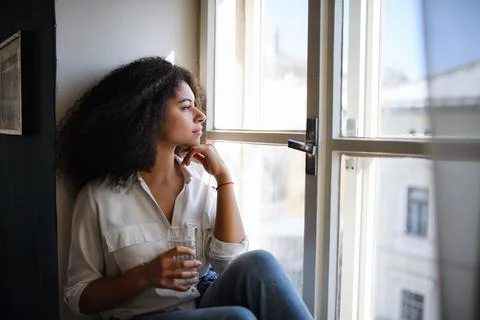 Portrait of young woman sitting on windowsill indoors, looking out and Stock Photos
