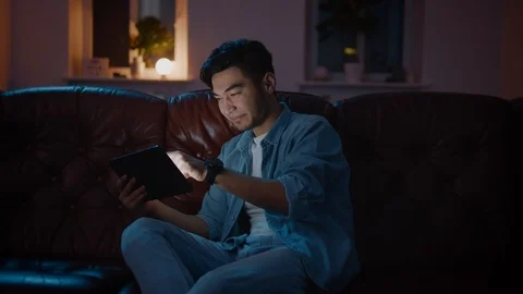 Positive Asian guy playing interactive game online on tablet home at night. Stockbeeldmateriaal 105585619