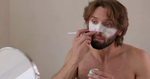 Positive bearded man using cosmetic brush for applying white clay mask on face Stock Footage 142528591