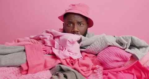 Positive dark skinned ethnic man wears panama covered with pile of laundry does Stock Footage 157336565