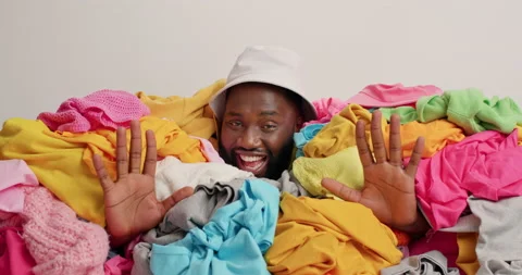 Positive dark skinned guy has cheerful expression sticks head in pile of laundry Stock Footage 157335464