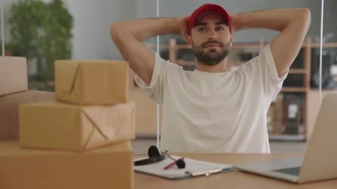 Positive delivery man using laptop computer near parcel boxes in office Stock Footage 153843654