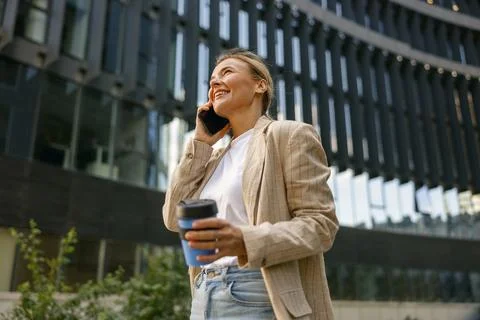 Positive female freelancer drinking coffee and talking phone standing on modern Stock Photos