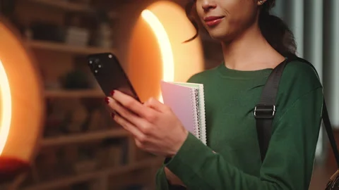 Positive female student scrolling through book reviews on phone in library Stock Footage 340961924