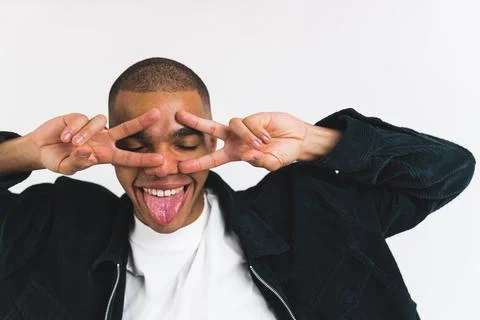 Positive funny male student posing with his tongue out and showing v sign with Stock Photos