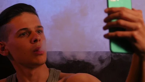 Positive guy doing selfie on smartphone with vapor from electronic cigarette at Video stock 106029849