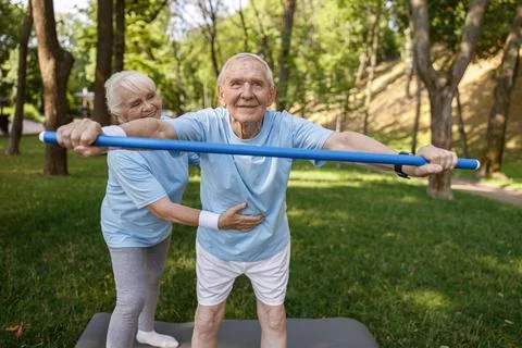 Positive lady helps senior man to do forward bend with bar in green park Stock Photos