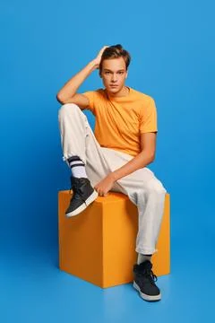 Positive man in orange undershirt and white sweatpants sits on wooden cube ov Foto stock