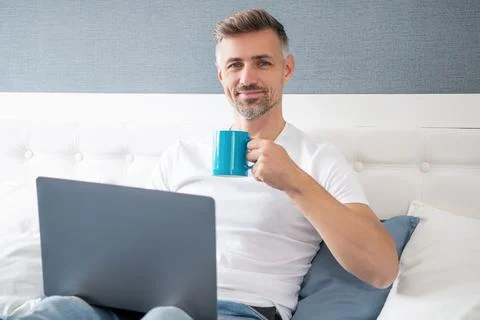 Positive mature man using computer in bed with coffee 스톡 사진
