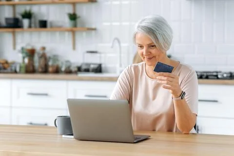 Positive middle aged caucasian gray haired female pensioner, sits at a table Stock Photos