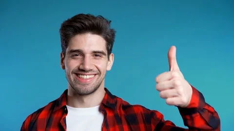 Positive young man smiles to camera. Hipster guy showing thumb up sign over blue Vidéo 120457097