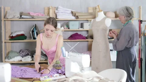Positive young tailoress cutting textile using scissors while other working with Video stock 346121956