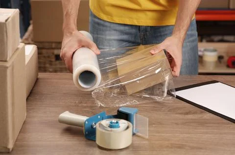 Post office worker wrapping parcel in stretch film at counter indoors, closeu Foto stock