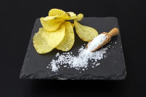 Potato chips stack nest to a scoop full of sea salt, on a black stone tile Stock Photos