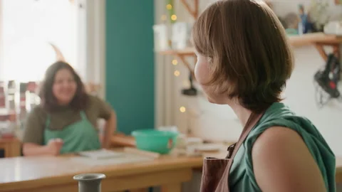 Potter and student sharing laugh at wheel, warm studio light, jars and tools on Stock Footage 346229085
