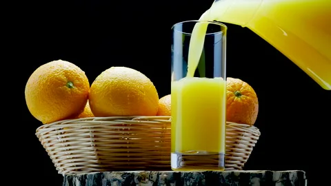 Pour orange juice into a glass on a black background. Fresh oranges in a basket Stock Footage