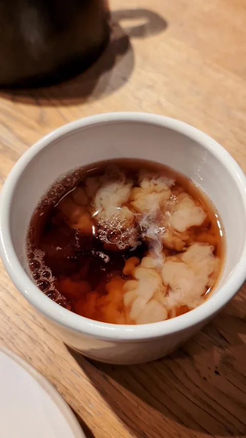 Pouring cream into black tea cup creating swirl on wooden cafe table Video stock 335636309