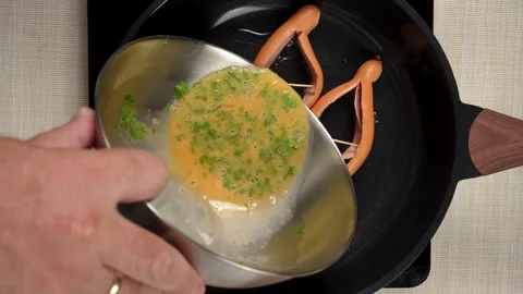 Pouring Egg and Parsley Mixture toward Split Sausages in a Pan Видео 343301419