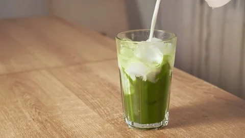 Pouring fresh milk over ice cubes into green matcha tea in tall glass. Stock Footage 345681689