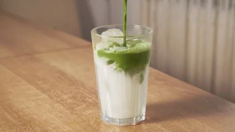 Pouring green matcha tea over iced milk in tall clear glass on wooden table Stock Footage 345262079