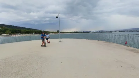 POV camera passing by two boys standing with scooters on a seaside promenade Stock Footage 318161441