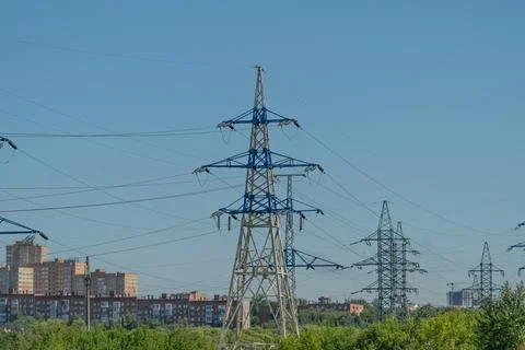 Power lines stand tall against a blue sky with a few clouds. Buildings are vi Stock Photos