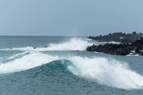Powerful ocean waves crash against black volcanic rocks under a blue sky Stock Photos