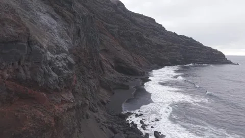 Powerful ocean waves pounding black volcanic beach, cascading against steep Stock Footage 308403580