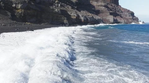 Powerful white waves break against dark volcanic sand, red cliff backdrop 스톡 동영상 308399866