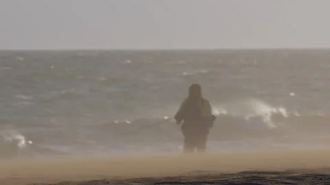 Powerful wind sweeping sand across barren coastal terrain, solitary fisher Stock Footage 306713733