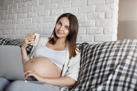 Pregnant lady using a laptop computer home sitting on sofa. Spending her free Stock Photos