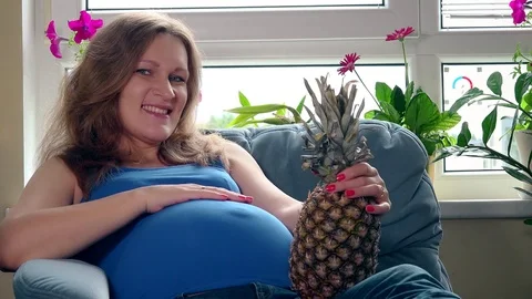 Pregnant woman with big belly holding pineapple ananas fruit sitting on sofa Stock Footage