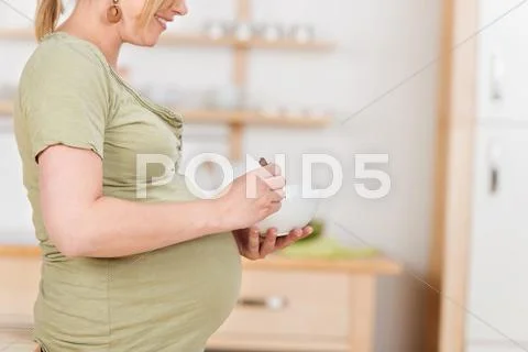 Pregnant woman eating muesli while sitting on kitchen counter Stock Photos