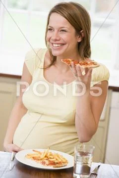 Pregnant woman in kitchen eating French fries and pizza smiling Stock Photos