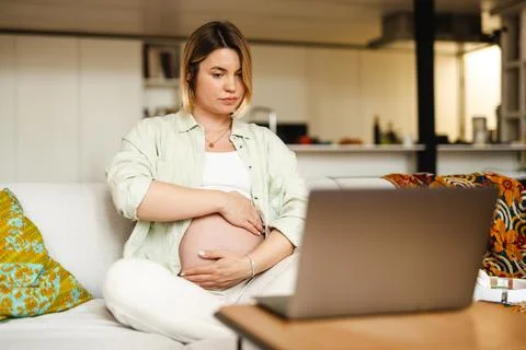 Pregnant woman showing her belly and using laptop while sitting on couch Stock Photos