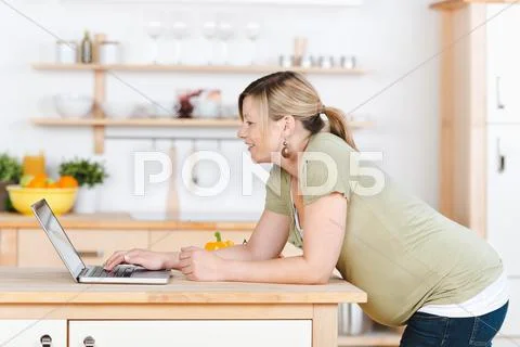 Pregnant woman using laptop while leaning on kitchen counter Stock Photos