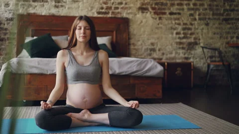 Pregnant young woman is sitting in lotus pose on yoga mat with palms on her Stock Footage