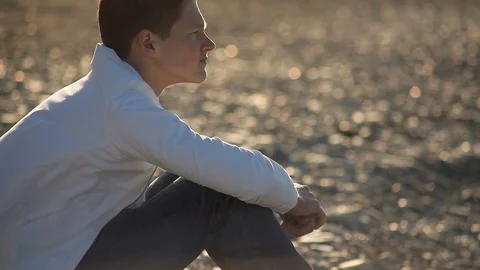 Preoccupied 16 year old boy sitting on the beach in the wind with his head on hi Stock Footage 114160275