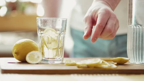 Preparing Fresh Lemon Water: Man's Hand Dropping Fruit and Pouring Mineral Water Stock Footage 332680674