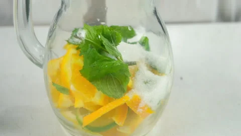 Preparing lemonade putting sugar to herb mint, citrus slices into glass jug. Stock Footage 313118223