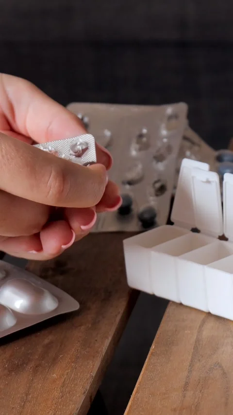 Pressing tablets from blister pack into hand with weekly pill organizer nearby Stock Footage 313794243