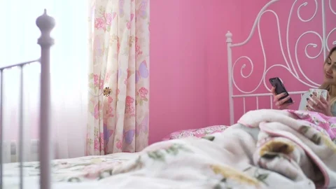 Pretty blonde lying in bed, drinking and using mobile phone in 4K 動画素材 72634891