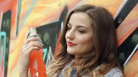 Pretty brunette looks happy while leaning on the graffiti wall and drinking juic Stock-Footage 79362530